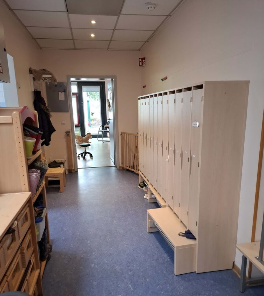 Ein Stück Flur bei dem auf der linken Seite offene Regale sind und rechts geschlossene Spinde in hellem Holz. Der Boden ist blau. Der Weg führt zur Marienkäfergruppe.
