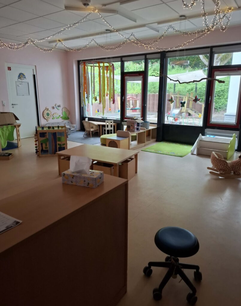 Es ist ein Zimmer mit Möbeln in hellem Holz zu sehen und Spielsachen. Vorne ist eine große Fensterfront mit Blick auf den Außenspielbereich.